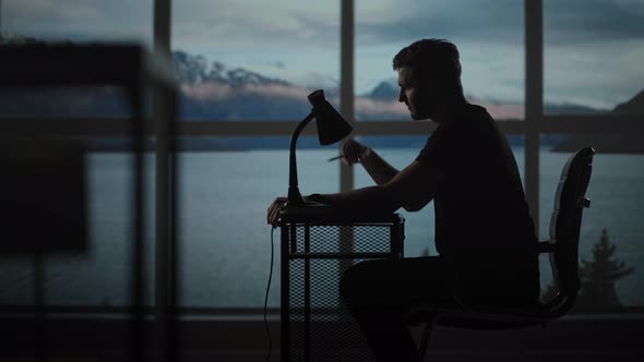 A Thoughtful Man Sitting at a Table with a Desk Lamp Writes with a Pen alt