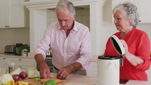 Happy caucasian senior couple in kitchen preparing meal, composting vegetable scraps alt