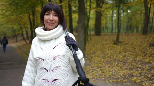 Smiling Caucasian Mature Woman Looking at Camera Standing in Autumn Park with Ski Poles alt