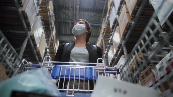 Woman in medical mask walking in mall steering shopping trolley looking around at shelves with goods alt