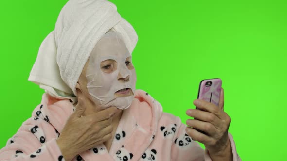 Grandmother in Bathrobe, Face Mask. Old Woman Using Mobile Phone for Video Call alt