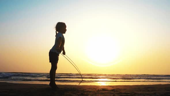 Little Girl Is Jumping on the Rope on the Sea Sand Beach at Sunset.