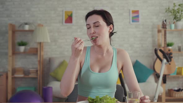 Happy and Joyful Young Sporty Woman Eating Vegetables Sitting at the Table alt