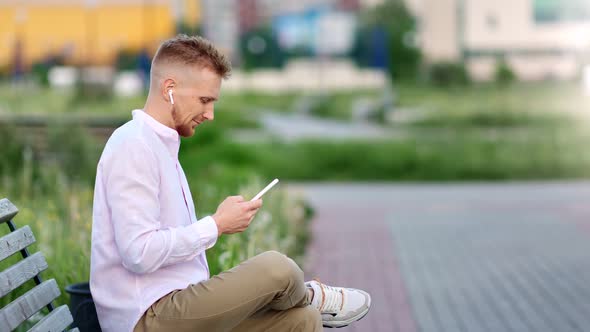 Attractive Hipster Male in Earphones Relaxing Sitting on Bench Listening Music Using Smartphone alt