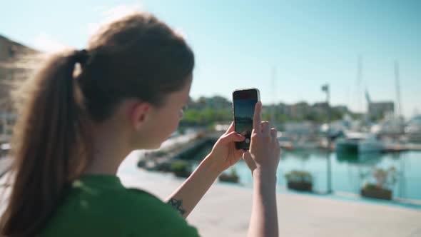 Blonde woman wearing green t-shirt making photo of the seafront on mobile near yachts alt