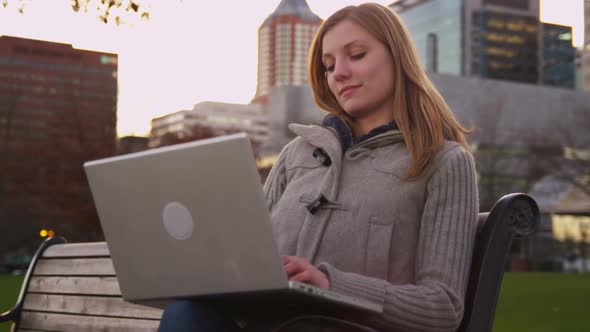 Young woman sits on park bench using laptop computer alt