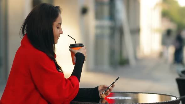 Fashionable Attractive Woman Drinks Coffee and Uses a Smartphone Sitting at a Table in a Cafe