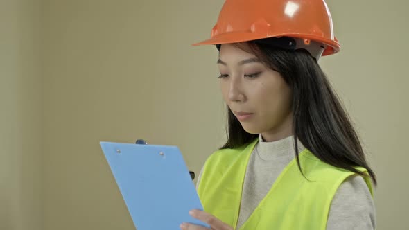 Asian Builder Woman Wearing Protective Clothing and a Helmet is Writing Down Something alt