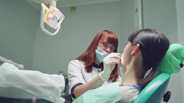 Red-haired female dentist looking at patient's teeth before treating them in stomatology office. alt