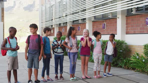 Diverse group of schoolchildren wearing backpacks smiling and standing in a row at school yard alt