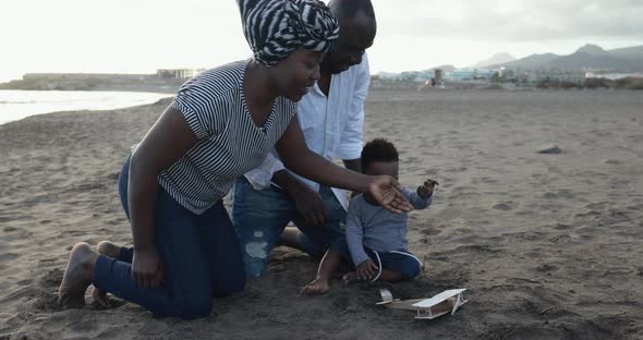 African parents and little son having fun with wood airplane on the beach alt