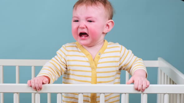 Cry infant baby boy stands in the crib, studio blue background. Sad child in yellow pajamas