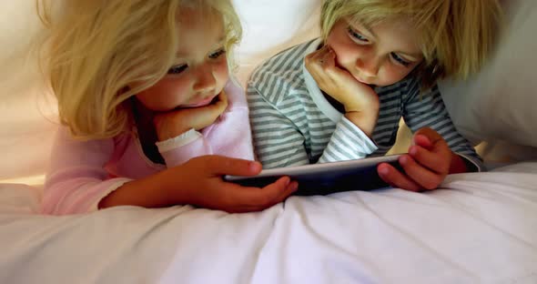 Siblings using digital tablet under blanket in bedroom at home 4k