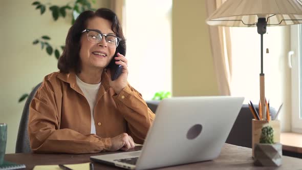Senior Woman Employee Talk on Phone When Working with Laptop alt