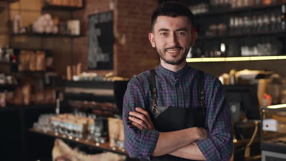 Portrait of Caucasian Barista Guy Wearing Apron Smiling and Standing with Arms Crossed Inside Bar or alt