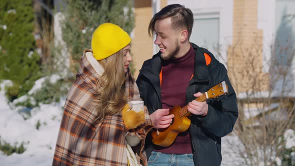 Young Man Playing Ukulele Singing Serenade for Smiling Woman Holding Coffee Tea Cup Outdoors on alt