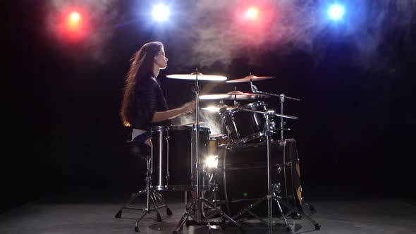 Girl Plays the Drums and Smiles. Black Smoke Background. Red Blue Light From Behind. Side View alt