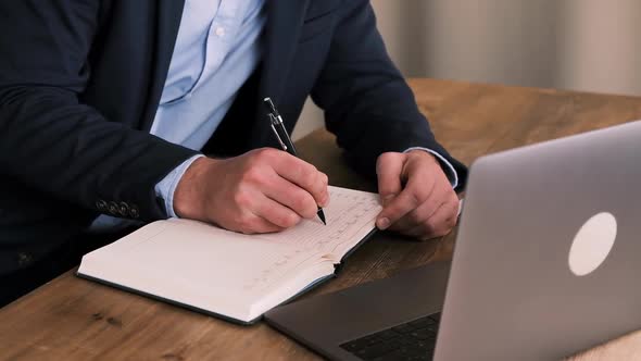 Cropped Side View of Business Man in Suit Writing Something on Notebook with an Open Laptop in the