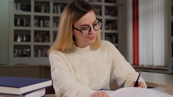 A Student Girl with Glasses Is Sitting at a Library Table with Books and Writing alt