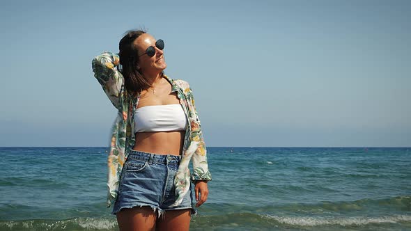 Cheerful carefree woman in summer tropical shirt and jeans shorts standing on seashore alt