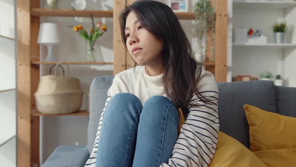 Asia lady sit embracing knees at sofa in living room at house look outside with feel lonely. alt