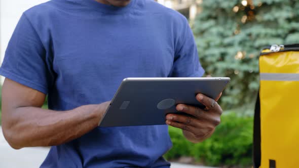 Close Up Afroamerican Man Courier Food Delivery Sitting on Bench Uses Tablet alt