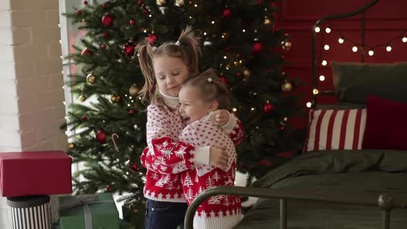 Two Little Caucasian Sisters Hug Together Near the Big Christmas Tree