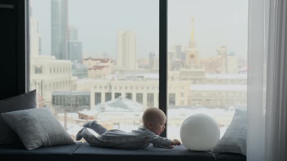 baby boy lies on the windowsill and looks out the window at houses