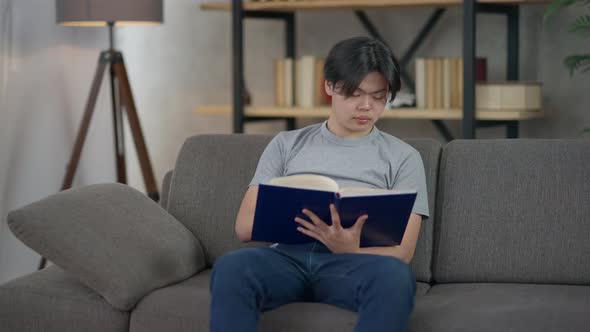 Young Asian Man Sitting on Couch in Living Room Turning Pages of Sketchpad alt
