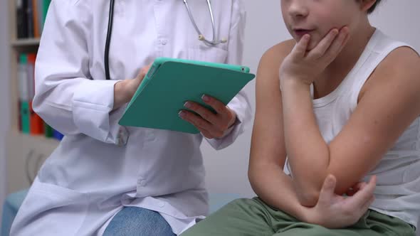 Unrecognizable Caucasian Doctor and Boy Sitting in Hospital with Tablet Talking alt