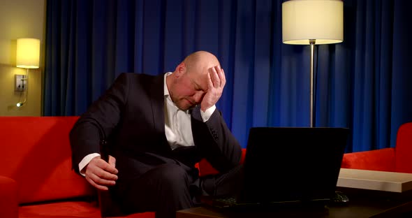 a Bald Man in a Dark Suit Is Sitting on a Red Sofa in Front of a Laptop Against a Blue Curtain. He alt