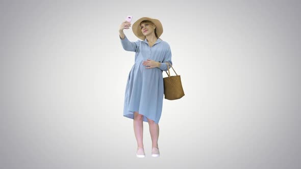 Happy Pregnant Woman in a Hat Using Her Smartphone Doing Selfie on Gradient Background alt