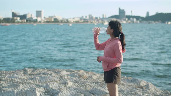 Asian woman runner drinking water while standing seaside. alt