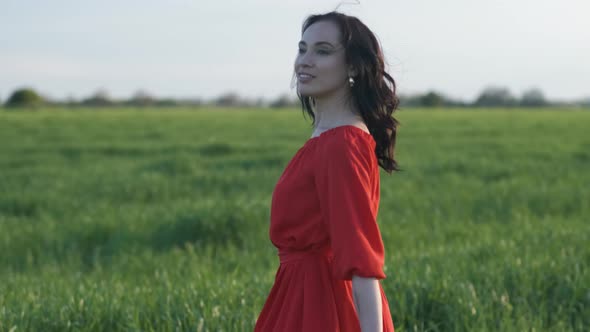 Beautiful Young Woman in a Red Dress Walks in a Green Wheat Field at Sunset or Dawn alt