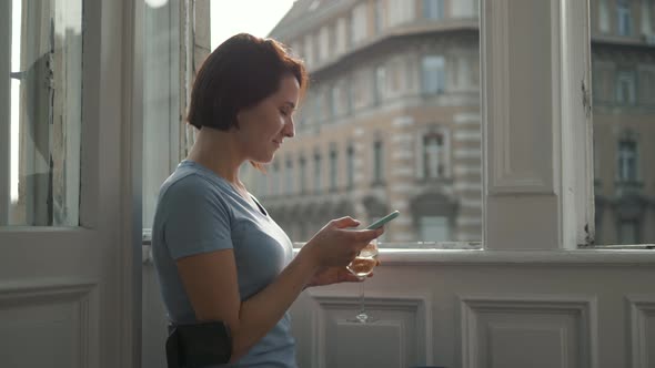 Side View of Young Woman Uses a Smartphone While Drinking Wine and Sitting By the Window at Home on alt
