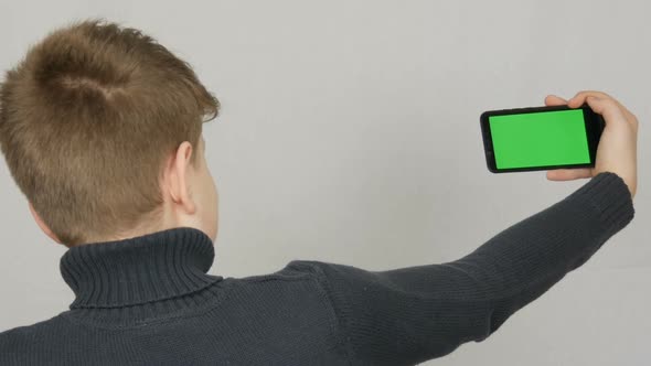 A Teen Boy Stands with His Back to the Camera and is Holding in Hand a Black Smartphone with a Green