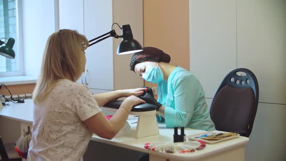 A manicurist in a protective mask doing a manicure to a client.