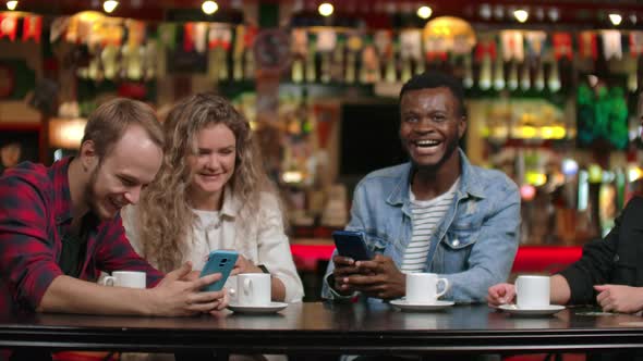 Four Friends Sitting in a Cafe Laugh and Smile While Looking at the Screens of Phones and Exchanging alt