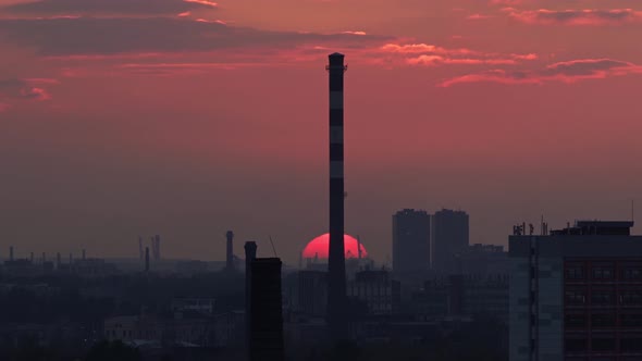 Red Sun Sinks Below the Horizon Against the Backdrop of the City's Industrial District Behind a Tall