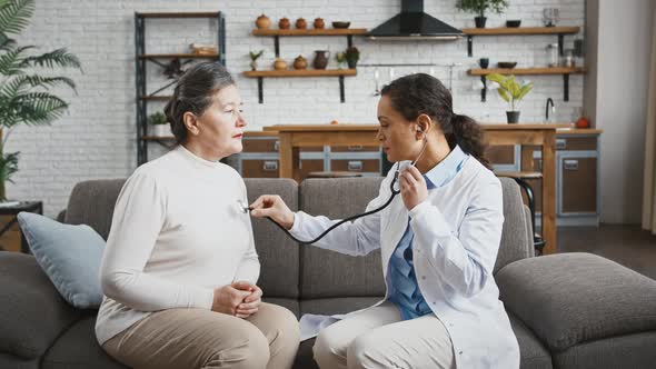 Woman Doctor Examining Aged Lady By Stethoscope While She is Breathing Deeply Sitting on Couch alt