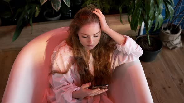 Pretty Girl With Long Hair In White Men's Shirt Sits In The Bathtub In The Bathroom alt