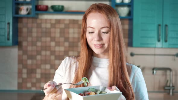 Attractive red-haired woman in kitchen holds plate with prepared vegetable salad alt