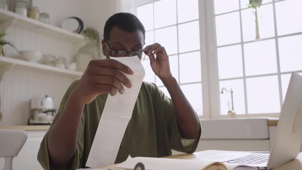 African American Man Looks at Checks Near Laptop in Kitchen alt