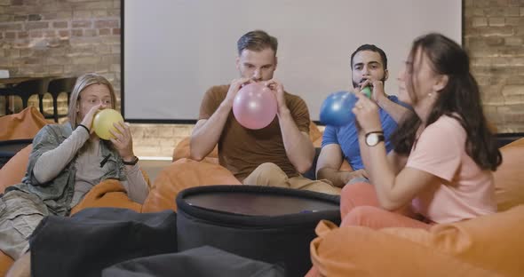 Relaxed Multiethnic Young Friends Sitting on Bag Chairs and Blowing Balloons. Portrait of Cheerful alt