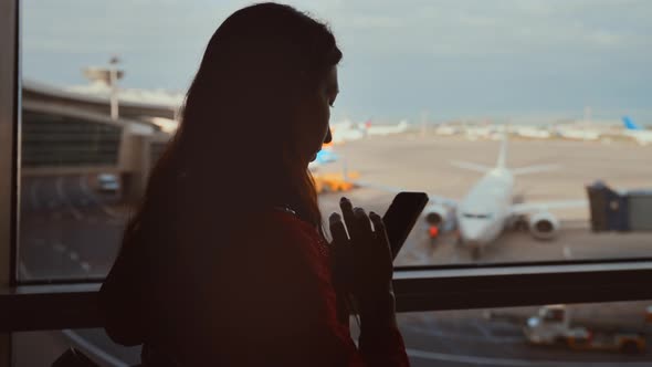 Businesswoman at Airport with Phone in Hands Against the Large Panoramic Window with Parking alt