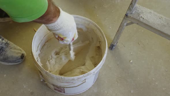 A man's gloved hand with a trowel stirring plaster in a bucket alt