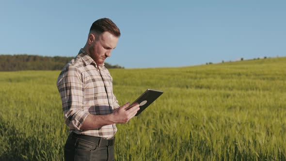 The Farmer is Standing Near the Field and Working with a Tablet alt