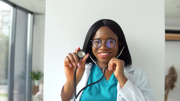 African American Woman Doctor in White Coat in Clinic at Workplace Holds Stethoscope in His Hand and alt
