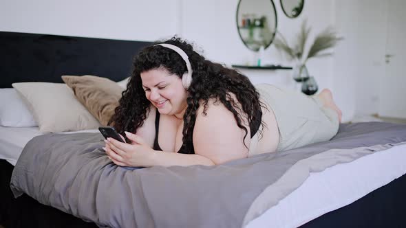 Young Overweight Brunette Lies on Bed Typing on Phone alt