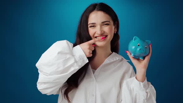 Pleased Young Woman Holding Piggy Bank Full of Money Against Blue Background